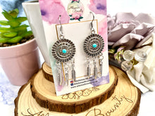 Load image into Gallery viewer, A pair of silver-colored THALIA Dreamcatcher Earrings with turquoise accents displayed on a card, with a blurred background suggesting a soft focus on the earrings.