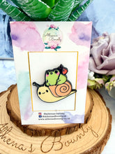 Load image into Gallery viewer, Pin with a design of a frog on a snail, displayed on a card with 'Athena's Bounty' branding, surrounded by plants.
